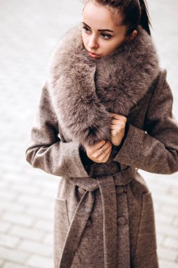 Açık havada ceketli bir kız. Dış giyim mankeni. Uzun bej ceketli esmer, kürklü, şehirde dolaşıyor. Şık bir manto içinde güzel bir kız. bahar sonbahar koleksiyonu.