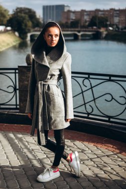 Açık havada ceketli bir kız. Dış giyim mankeni. Uzun gri paltolu bir esmer nehrin seti boyunca yürüyor. Şık bir manto içinde güzel bir kız. bahar sonbahar koleksiyonu.