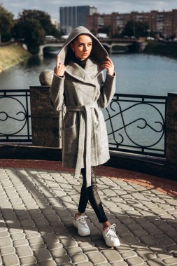 Açık havada ceketli bir kız. Dış giyim mankeni. Uzun gri paltolu bir esmer nehrin seti boyunca yürüyor. Şık bir manto içinde güzel bir kız. bahar sonbahar koleksiyonu.