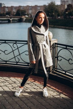 Açık havada ceketli bir kız. Dış giyim mankeni. Uzun gri paltolu bir esmer nehrin seti boyunca yürüyor. Şık bir manto içinde güzel bir kız. bahar sonbahar koleksiyonu.