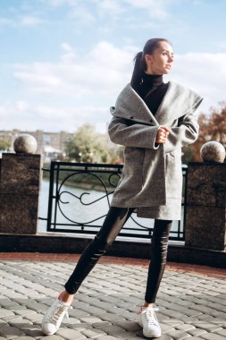 Açık havada ceketli bir kız. Dış giyim mankeni. Uzun gri paltolu bir esmer nehrin seti boyunca yürüyor. Şık bir manto içinde güzel bir kız. bahar sonbahar koleksiyonu.