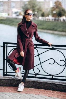 Açık havada ceketli bir kız. Dış giyim mankeni. Uzun bordo paltolu bir esmer nehrin seti boyunca yürür. Şık bir manto içinde güzel bir kız. bahar sonbahar koleksiyonu.