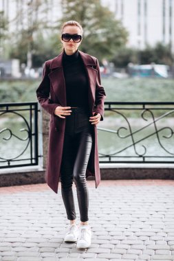 Açık havada ceketli bir kız. Dış giyim mankeni. Uzun bordo paltolu bir esmer nehrin seti boyunca yürür. Şık bir manto içinde güzel bir kız. bahar sonbahar koleksiyonu.