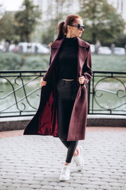 Açık havada ceketli bir kız. Dış giyim mankeni. Uzun bordo paltolu bir esmer nehrin seti boyunca yürür. Şık bir manto içinde güzel bir kız. bahar sonbahar koleksiyonu.