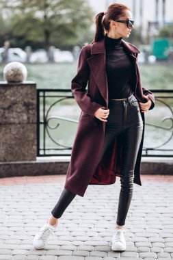 Açık havada ceketli bir kız. Dış giyim mankeni. Uzun bordo paltolu bir esmer nehrin seti boyunca yürür. Şık bir manto içinde güzel bir kız. bahar sonbahar koleksiyonu.
