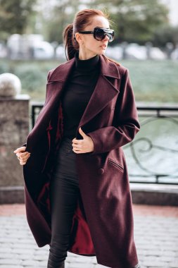 Açık havada ceketli bir kız. Dış giyim mankeni. Uzun bordo paltolu bir esmer nehrin seti boyunca yürür. Şık bir manto içinde güzel bir kız. bahar sonbahar koleksiyonu.