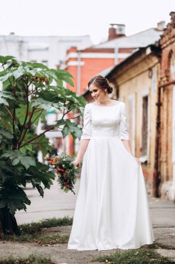 Mütevazı bir elbise içindeki güzel bir gelinin portresi. Uzun kollu, beyaz elbiseli esmer kız. Sonbahar buketi olan tatlı bir gelin. Bir sonbahar sokağında açık havada bir kadının portresi..
