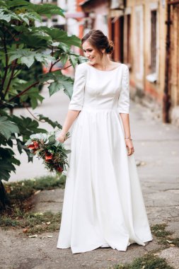 Mütevazı bir elbise içindeki güzel bir gelinin portresi. Uzun kollu, beyaz elbiseli esmer kız. Sonbahar buketi olan tatlı bir gelin. Bir sonbahar sokağında açık havada bir kadının portresi..