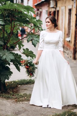 Mütevazı bir elbise içindeki güzel bir gelinin portresi. Uzun kollu, beyaz elbiseli esmer kız. Sonbahar buketi olan tatlı bir gelin. Bir sonbahar sokağında açık havada bir kadının portresi..