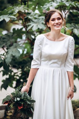 Mütevazı bir elbise içindeki güzel bir gelinin portresi. Uzun kollu, beyaz elbiseli esmer kız. Sonbahar buketi olan tatlı bir gelin. Bir sonbahar sokağında açık havada bir kadının portresi..