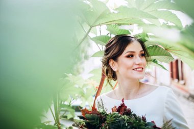 Mütevazı bir elbise içindeki güzel bir gelinin portresi. Uzun kollu, beyaz elbiseli esmer kız. Sonbahar buketi olan tatlı bir gelin. Güz sokağında açık havada çekilmiş bir kadın portresi..