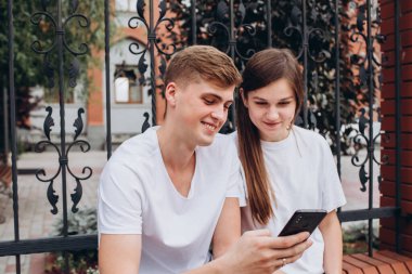 Genç bir çift karton bardaklardan kahve içer ve akıllı telefonlara bakar. Genç erkekler ve kızlar dışarıda oturuyorlar. Modern teknolojiler. Modern toplumda iletişim sorunu.