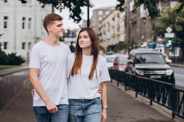 Genç çift şehrin sokaklarında yürüyor ve el ele tutuşuyorlar. Beyaz tişörtlü erkek ve kız açık havada kot pantolon giyiyor. Gençler sonbahar şehrinde yürürler. Mutlu genç çift.