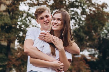 Mutlu genç çift şehrin sokaklarında yürüyor ve el ele tutuşuyorlar. Beyaz tişörtlü erkek ve kız açık havada kot pantolon giyiyor. Gençler bir sonbahar ağacının arka planına sarılıyorlar. Çift portresi.