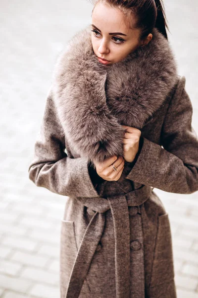 Açık havada ceketli bir kız. Dış giyim mankeni. Uzun bej ceketli esmer, kürklü, şehirde dolaşıyor. Şık bir manto içinde güzel bir kız. bahar sonbahar koleksiyonu.