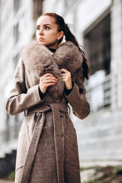 Açık havada ceketli bir kız. Dış giyim mankeni. Uzun bej ceketli esmer, kürklü, şehirde dolaşıyor. Şık bir manto içinde güzel bir kız. bahar sonbahar koleksiyonu.