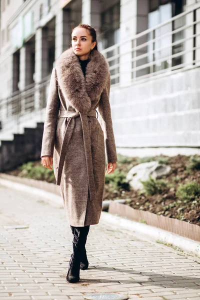 Açık havada ceketli bir kız. Dış giyim mankeni. Uzun bej ceketli esmer, kürklü, şehirde dolaşıyor. Şık bir manto içinde güzel bir kız. bahar sonbahar koleksiyonu.