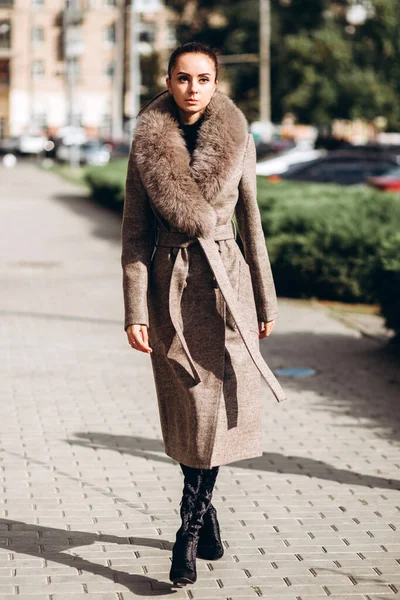 Açık havada ceketli bir kız. Dış giyim mankeni. Uzun bej ceketli esmer, kürklü, şehirde dolaşıyor. Şık bir manto içinde güzel bir kız. bahar sonbahar koleksiyonu.