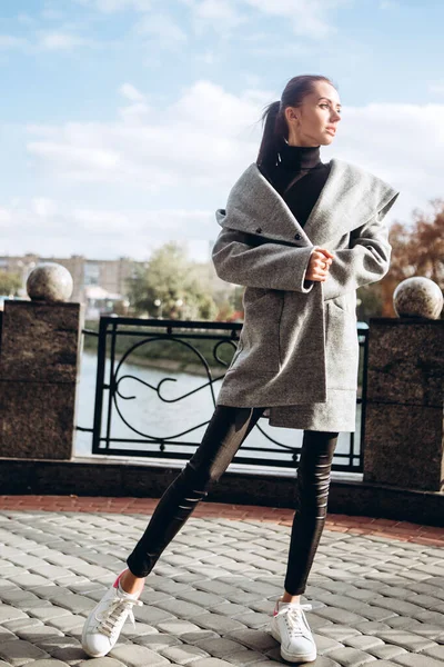 Açık havada ceketli bir kız. Dış giyim mankeni. Uzun gri paltolu bir esmer nehrin seti boyunca yürüyor. Şık bir manto içinde güzel bir kız. bahar sonbahar koleksiyonu.