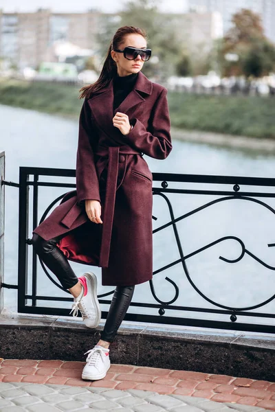 Açık havada ceketli bir kız. Dış giyim mankeni. Uzun bordo paltolu bir esmer nehrin seti boyunca yürür. Şık bir manto içinde güzel bir kız. bahar sonbahar koleksiyonu.
