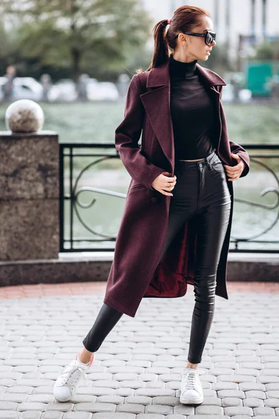 Açık havada ceketli bir kız. Dış giyim mankeni. Uzun bordo paltolu bir esmer nehrin seti boyunca yürür. Şık bir manto içinde güzel bir kız. bahar sonbahar koleksiyonu.