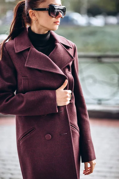 Açık havada ceketli bir kız. Dış giyim mankeni. Uzun bordo paltolu bir esmer nehrin seti boyunca yürür. Şık bir manto içinde güzel bir kız. bahar sonbahar koleksiyonu.