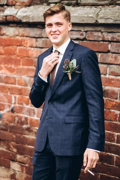 brutal groom in a suit and tie smokes a cigarette. The blond guy is walking around the old town. smoking, bad habits. Young male businessman on brick wall background. Portert of a smiling guy.