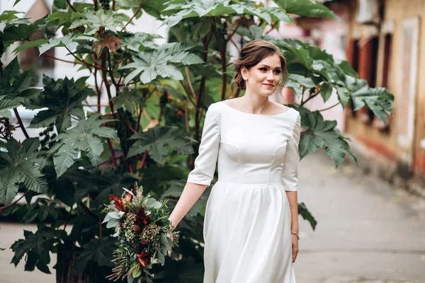 Portrait of a beautiful bride in a modest dress. Young brunette girl in a white dress with long sleeves. Cute bride with an autumn bouquet. Portrait of a woman outdoors on an autumn street.