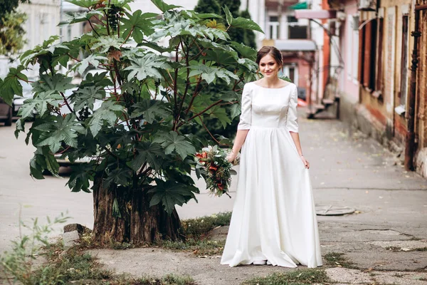 Portrait of a beautiful bride in a modest dress. Young brunette girl in a white dress with long sleeves. Cute bride with an autumn bouquet. Portrait of a woman outdoors on an autumn street.