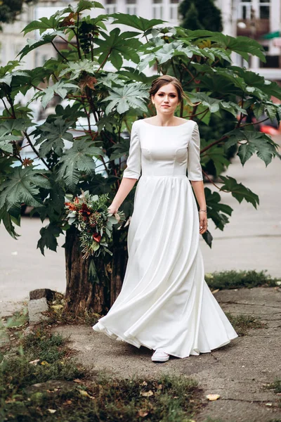 Portrait of a beautiful bride in a modest dress. Young brunette girl in a white dress with long sleeves. Cute bride with an autumn bouquet. Portrait of a woman outdoors on an autumn street.