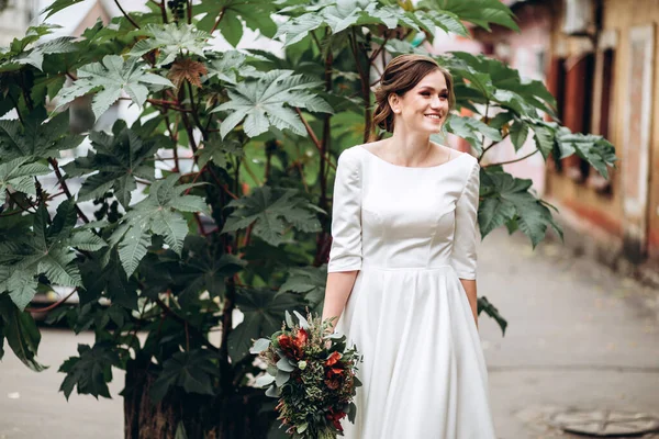Mütevazı bir elbise içindeki güzel bir gelinin portresi. Uzun kollu, beyaz elbiseli esmer kız. Sonbahar buketi olan tatlı bir gelin. Bir sonbahar sokağında açık havada bir kadının portresi..