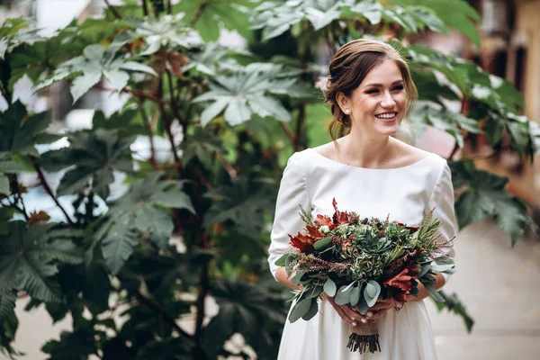 Mütevazı bir elbise içindeki güzel bir gelinin portresi. Uzun kollu, beyaz elbiseli esmer kız. Sonbahar buketi olan tatlı bir gelin. Bir sonbahar sokağında açık havada bir kadının portresi..