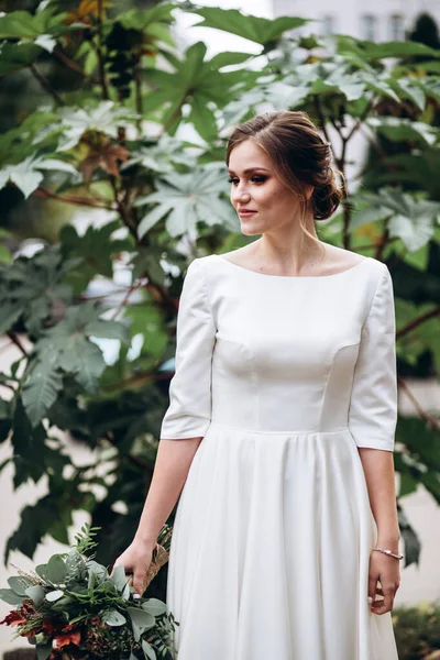 Mütevazı bir elbise içindeki güzel bir gelinin portresi. Uzun kollu, beyaz elbiseli esmer kız. Sonbahar buketi olan tatlı bir gelin. Bir sonbahar sokağında açık havada bir kadının portresi..