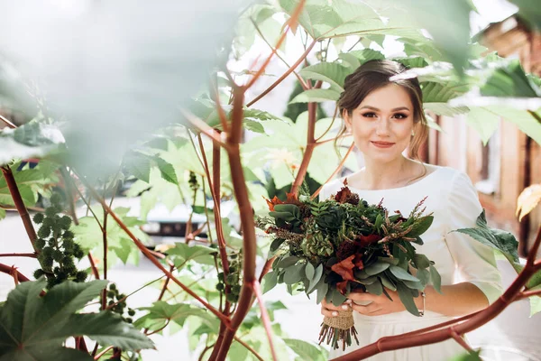 Mütevazı bir elbise içindeki güzel bir gelinin portresi. Uzun kollu, beyaz elbiseli esmer kız. Sonbahar buketi olan tatlı bir gelin. Güz sokağında açık havada çekilmiş bir kadın portresi..