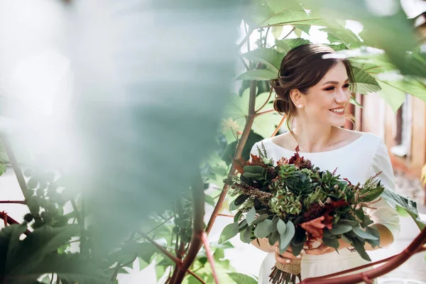 Mütevazı bir elbise içindeki güzel bir gelinin portresi. Uzun kollu, beyaz elbiseli esmer kız. Sonbahar buketi olan tatlı bir gelin. Güz sokağında açık havada çekilmiş bir kadın portresi..