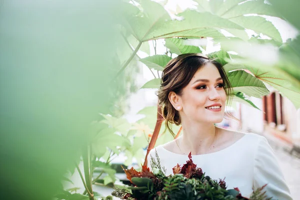 Mütevazı bir elbise içindeki güzel bir gelinin portresi. Uzun kollu, beyaz elbiseli esmer kız. Sonbahar buketi olan tatlı bir gelin. Güz sokağında açık havada çekilmiş bir kadın portresi..