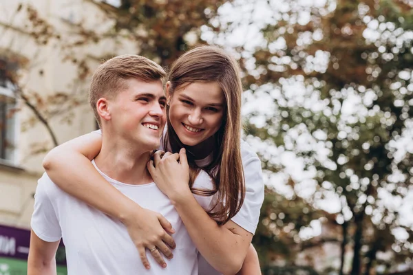 Mutlu genç çift şehrin sokaklarında yürüyor ve el ele tutuşuyorlar. Beyaz tişörtlü erkek ve kız açık havada kot pantolon giyiyor. Gençler bir sonbahar ağacının arka planına sarılıyorlar. Çift portresi.