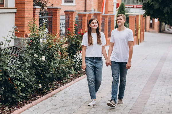 Mutlu genç çift şehrin sokaklarında yürüyor ve el ele tutuşuyorlar. Beyaz tişörtlü erkek ve kız açık havada kot pantolon giyiyor. Gençler sonbahar şehrinde duvarın arkasında yürüyorlar..
