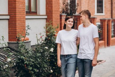 Mutlu genç çift şehrin sokaklarında yürüyor ve el ele tutuşuyorlar. Beyaz tişörtlü erkek ve kız açık havada kot pantolon giyiyor. Gençler sonbahar şehrinde duvarın arkasında yürüyorlar..