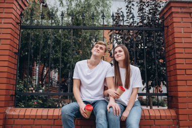 Genç çift karton bardaklardan kahve içer. Mutlu bir çift tuğla bir çitte oturuyor ve gülüyor. Gerçek duygular. Yakın plan bir çift portre. Erkek ve kadın güneşli bir günde sokakta yürürler..