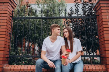 Genç çift karton bardaklardan kahve içer. Mutlu bir çift tuğla bir çitte oturuyor ve gülüyor. Gerçek duygular. Yakın plan bir çift portre. Erkek ve kadın güneşli bir günde sokakta yürürler..