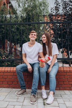 Genç çift karton bardaklardan kahve içer. Mutlu bir çift tuğla bir çitte oturuyor ve gülüyor. Gerçek duygular. Yakın plan bir çift portre. Erkek ve kadın güneşli bir günde sokakta yürürler..