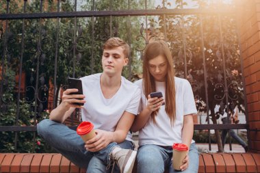 Genç bir çift karton bardaklardan kahve içer ve akıllı telefonlara bakar. Genç erkekler ve kızlar dışarıda oturuyorlar. Modern teknolojiler. Modern toplumda iletişim sorunu.