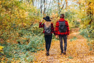 Genç çift sonbahar ormanında el ele tutuşarak yürüyor. Güzel giyimli bir erkek ve kız. Adam ve kırmızı ceketli kız sarı ormanın arka planında. Parkta aşık mutlu genç bir çift
