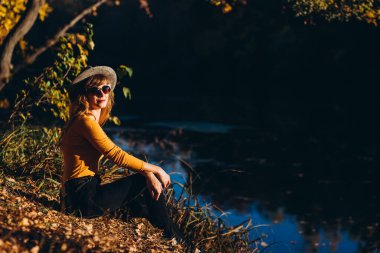 Gri şapkalı bir sarışın sonbahar ormanında yürüyor. Sarı kazaklı neşeli bir kız güneşli bir sonbahar gününde gölün kenarında oturur. Ormanda yürüyen bir kadın.