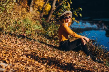 Gri şapkalı bir sarışın sonbahar ormanında yürüyor. Sarı kazaklı neşeli bir kız güneşli bir sonbahar gününde gölün kenarında oturur. Ormanda yürüyen bir kadın.