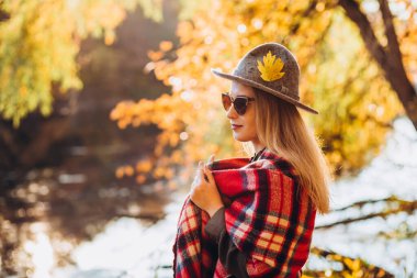 Gri şapkalı sarışın sonbahar ormanında yürüyor. Kırmızı kareli eşarplı neşeli kız sarı ormanın arka planında duruyor. Sonbaharda bir kızın yakın plan portresi. Bulanık arkaplan