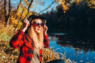 Gri şapkalı bir sarışın sonbahar ormanında yürüyor. Kırmızı ceketli neşeli kız güneşli bir sonbahar gününde gölde oturur. Ormanda yürüyen bir kadın.
