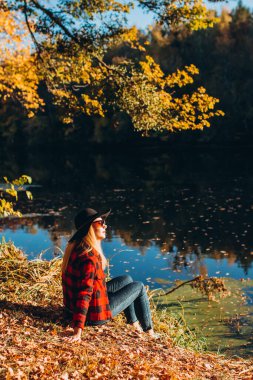 Gri şapkalı bir sarışın sonbahar ormanında yürüyor. Kırmızı ceketli neşeli kız güneşli bir sonbahar gününde gölde oturur. Ormanda yürüyen bir kadın.
