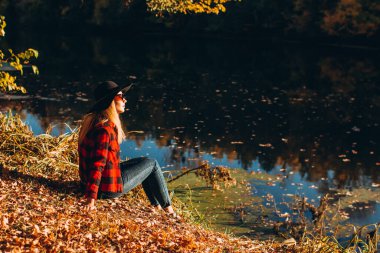Gri şapkalı bir sarışın sonbahar ormanında yürüyor. Kırmızı ceketli neşeli kız güneşli bir sonbahar gününde gölde oturur. Ormanda yürüyen bir kadın.